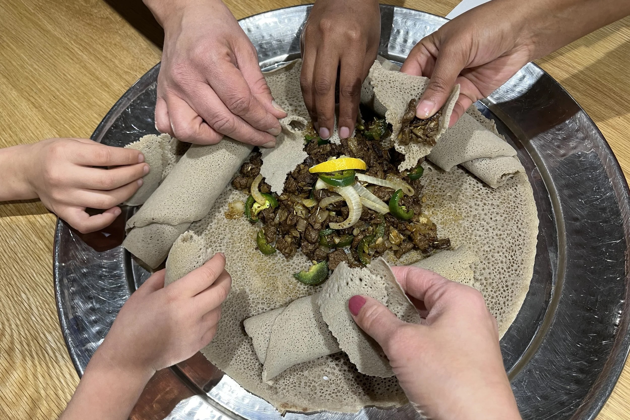 Multiple hands reaching for injera topped with seasoned slices of meat
