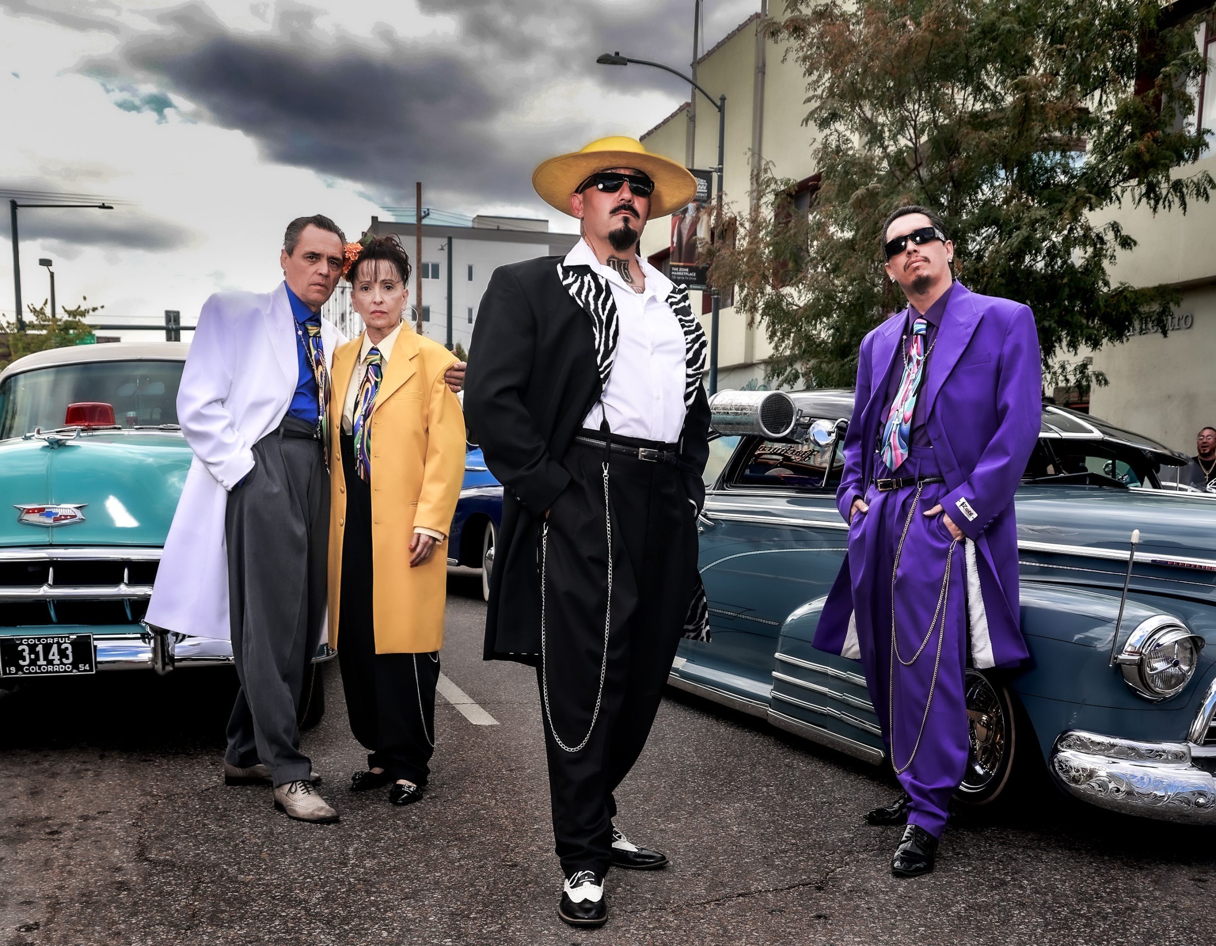 People dressed in zoot suits stand near low-rider cars