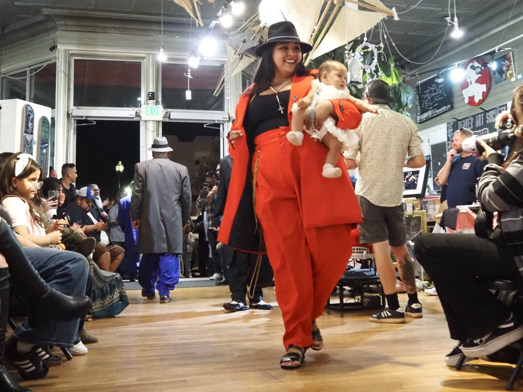 A woman in a red suit carrying a baby walks a runway