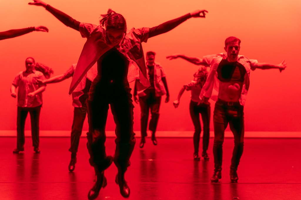 People dance onstage in orange light with their arms extended out.
