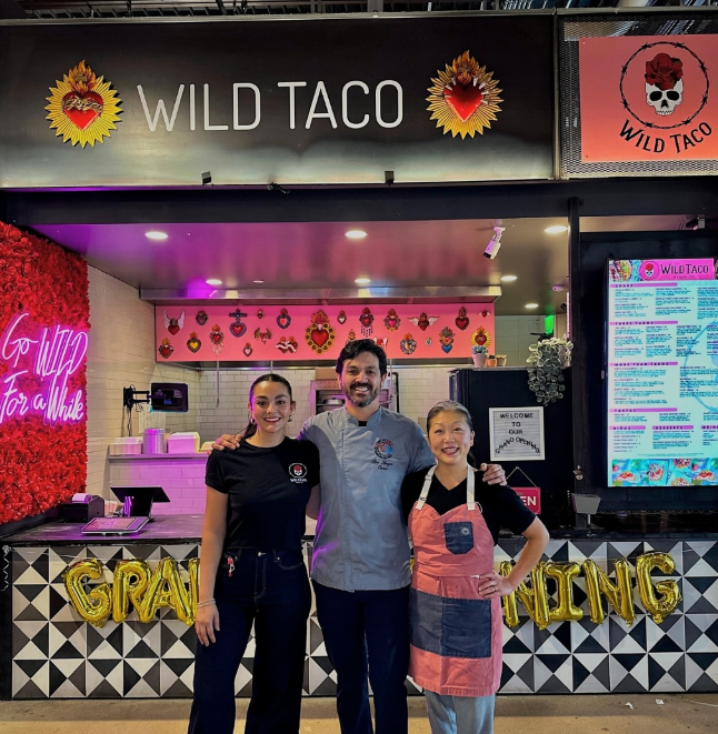 three people posing in front of a food hall stall