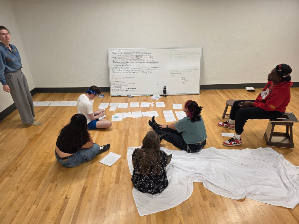 Theater artist gather around a white board with notes spread out around it.