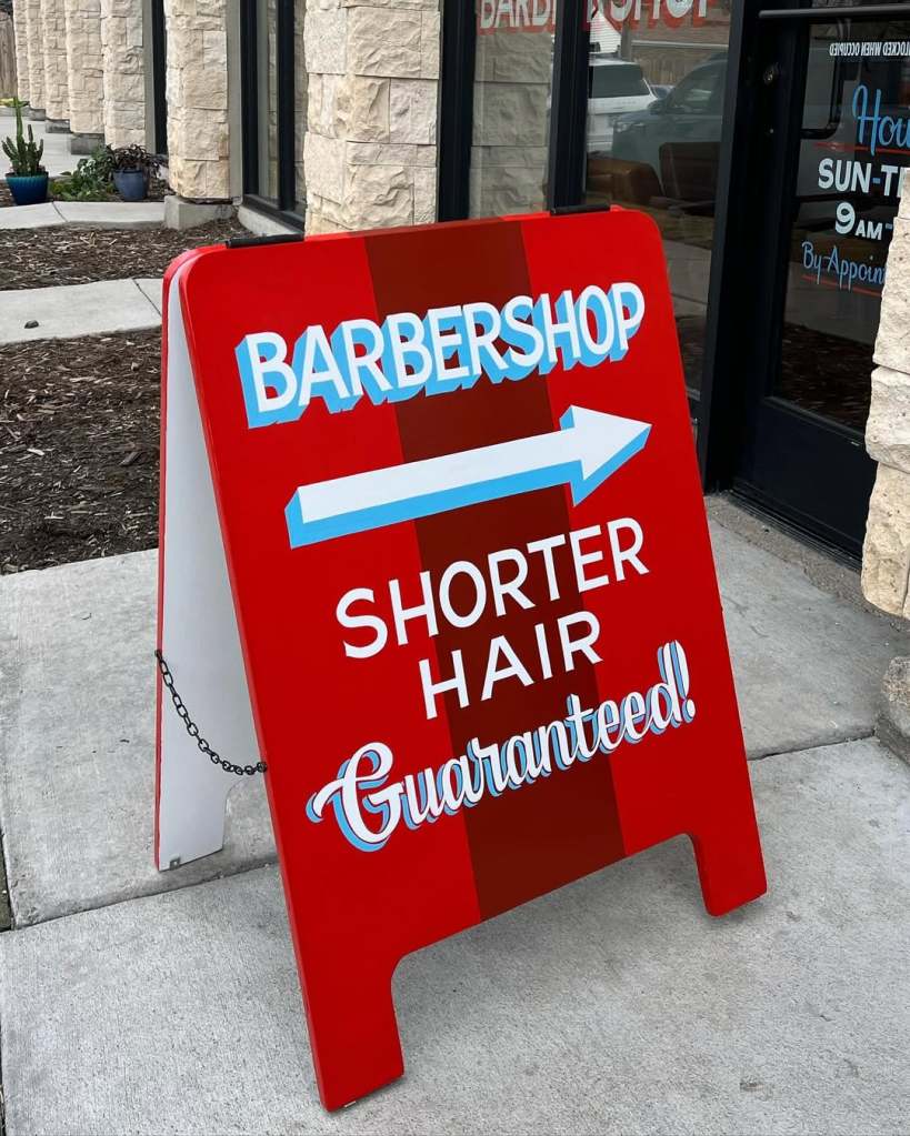 A-frame sign stacked top to bottom with "BARBERSHOP," the image of an arrow, and "SHORTER HAIR Guaranteed!"
