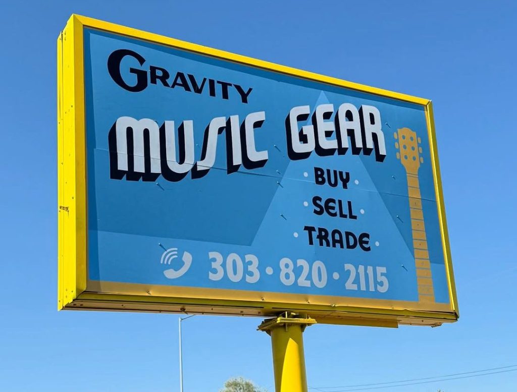 Outdoor sign captioned "GRAVITY MUSIC GEAR" with a list that says "BUY", "SELL", "TRADE", and the phone number 303-820-2115.