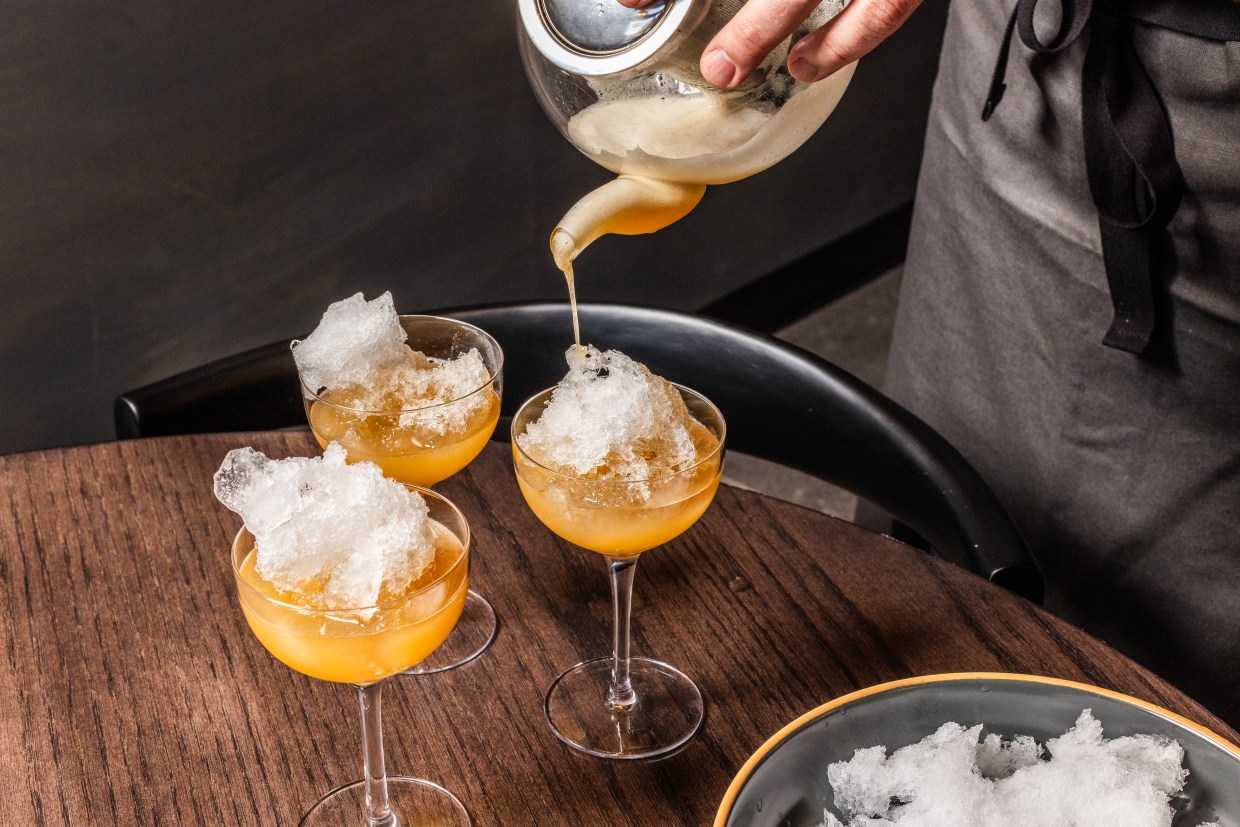 A peach-hued margarita pouring from a clear teapot into glasses of shaved ice