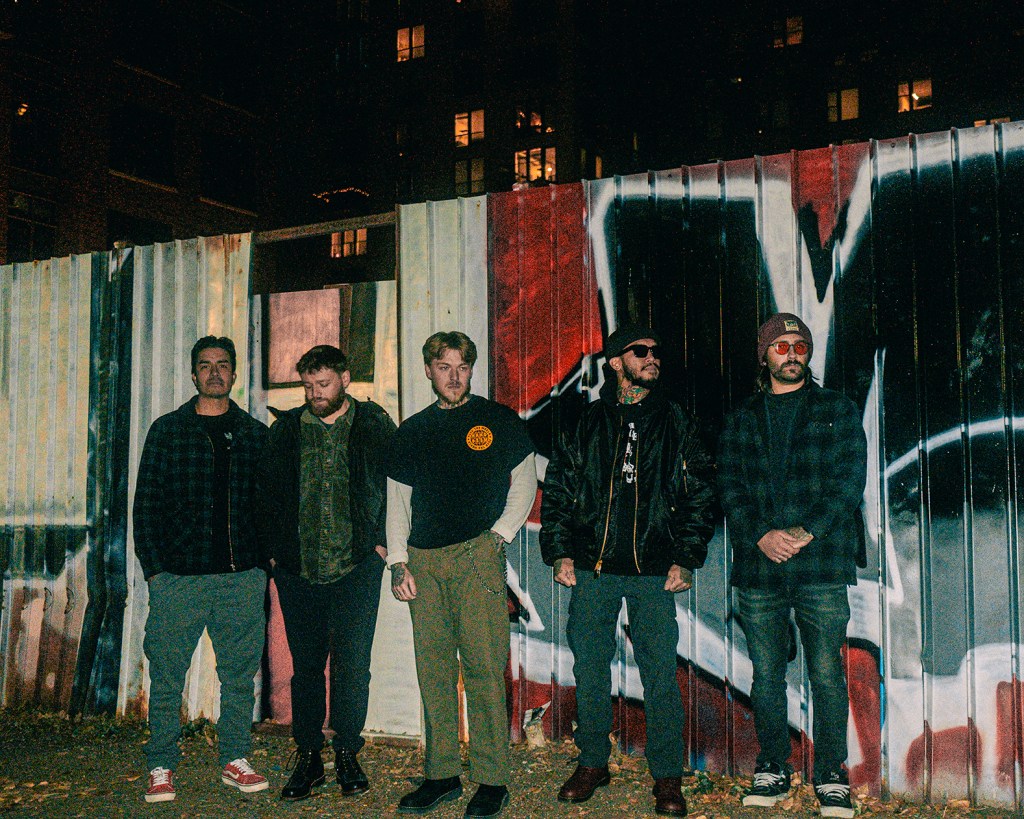 A photo of five band members in front of a wall with graffiti on it