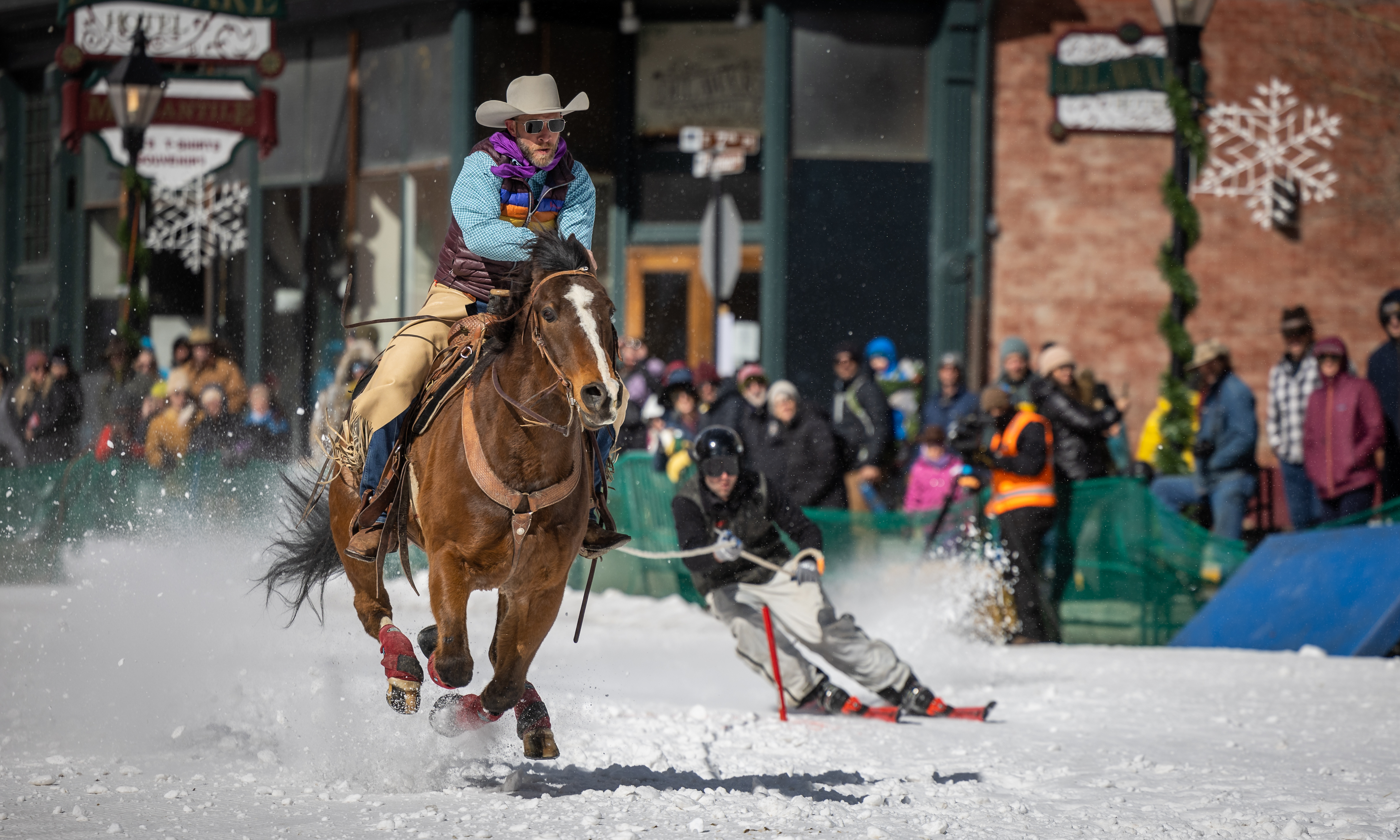 A person riding on horseback through downtown Leadville with a skier in tow