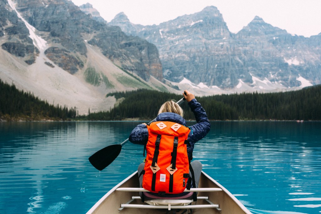 A person in a canoe, backdropped by mountains while wearing an orange Topo Designs pack