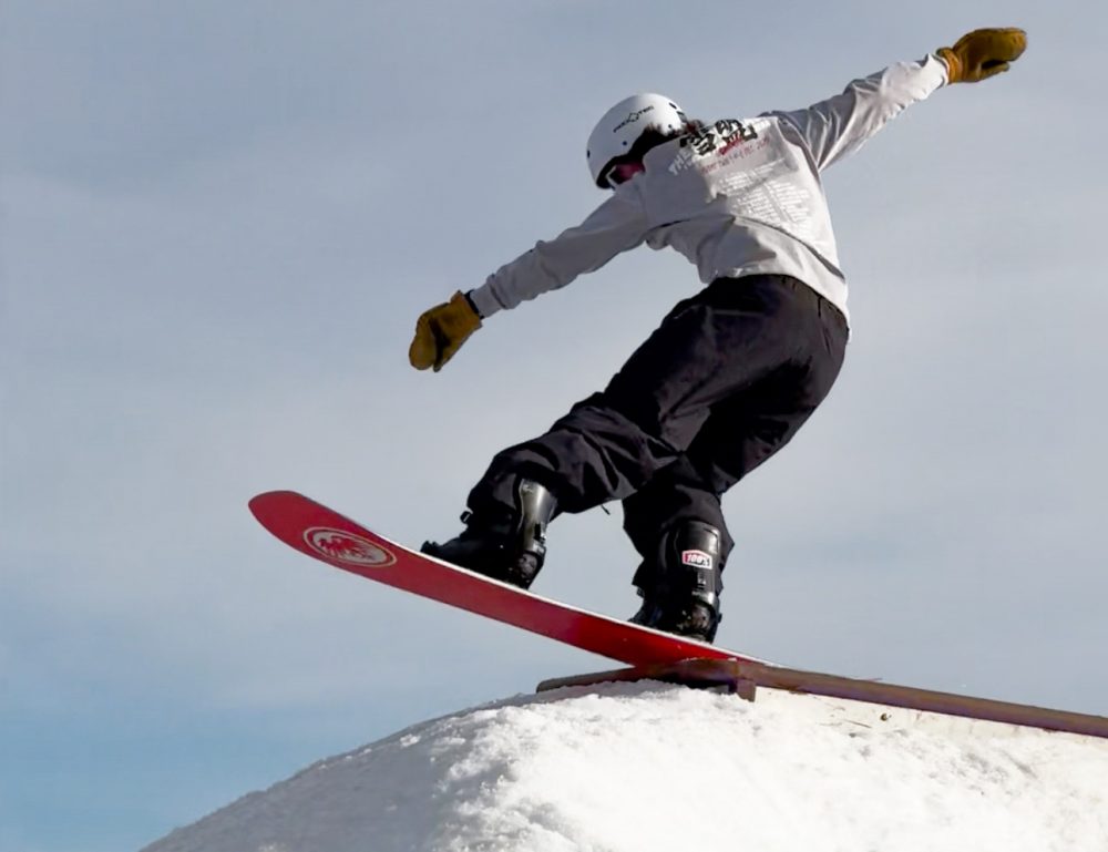 Chase Blackwell snowboarding at Ruby Hill Rail Yard. Blackwell has black snow pants and a board with a red bottom. He's about to jump off the top of the hill and perform a trick.