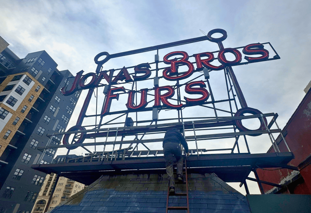 A man climbs a ladder up to the red "Jonas Bros Furs" neon sign atop a roof.