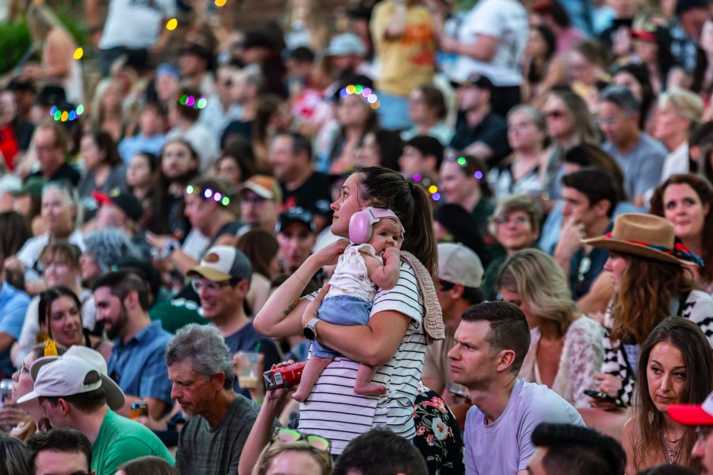 A photo showing a woman at an outdoor event with a baby wearing noise protecting headphones.