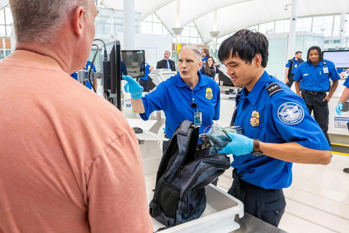 Customs officers question a traveler at an airport security checkpoint, inspecting his belongings and discussing security procedures.