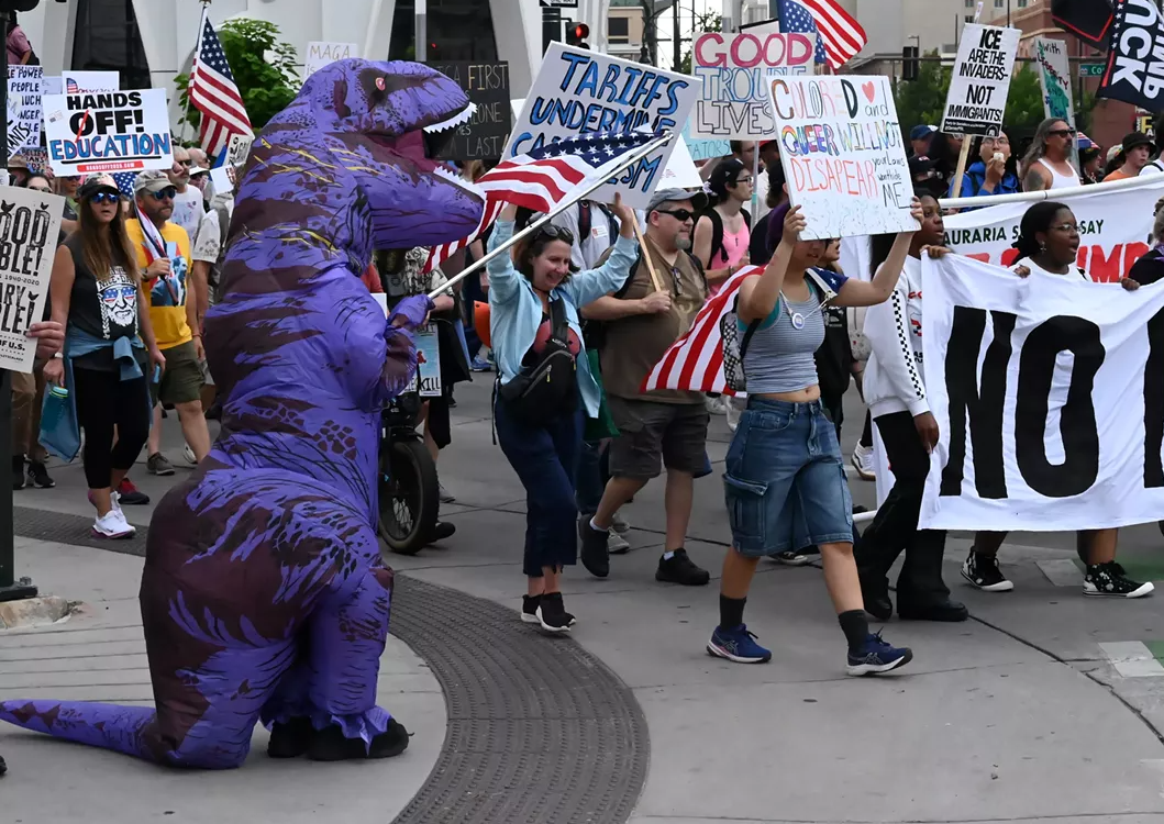 A person in a purple t-rex costume waves a flag as protesters walk down a street in downtown denver