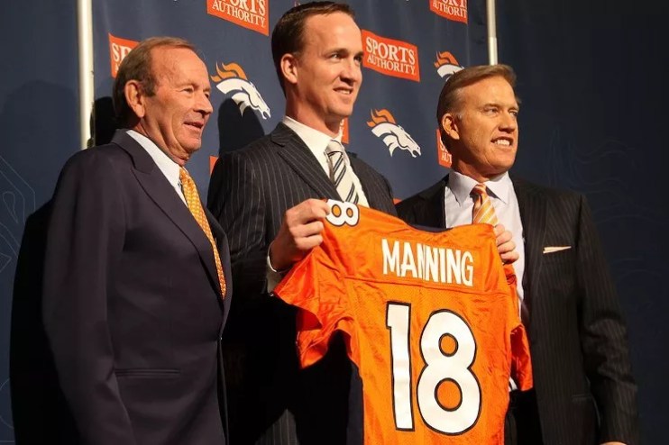 Three men stand in a row, one holding up an orange jersey.