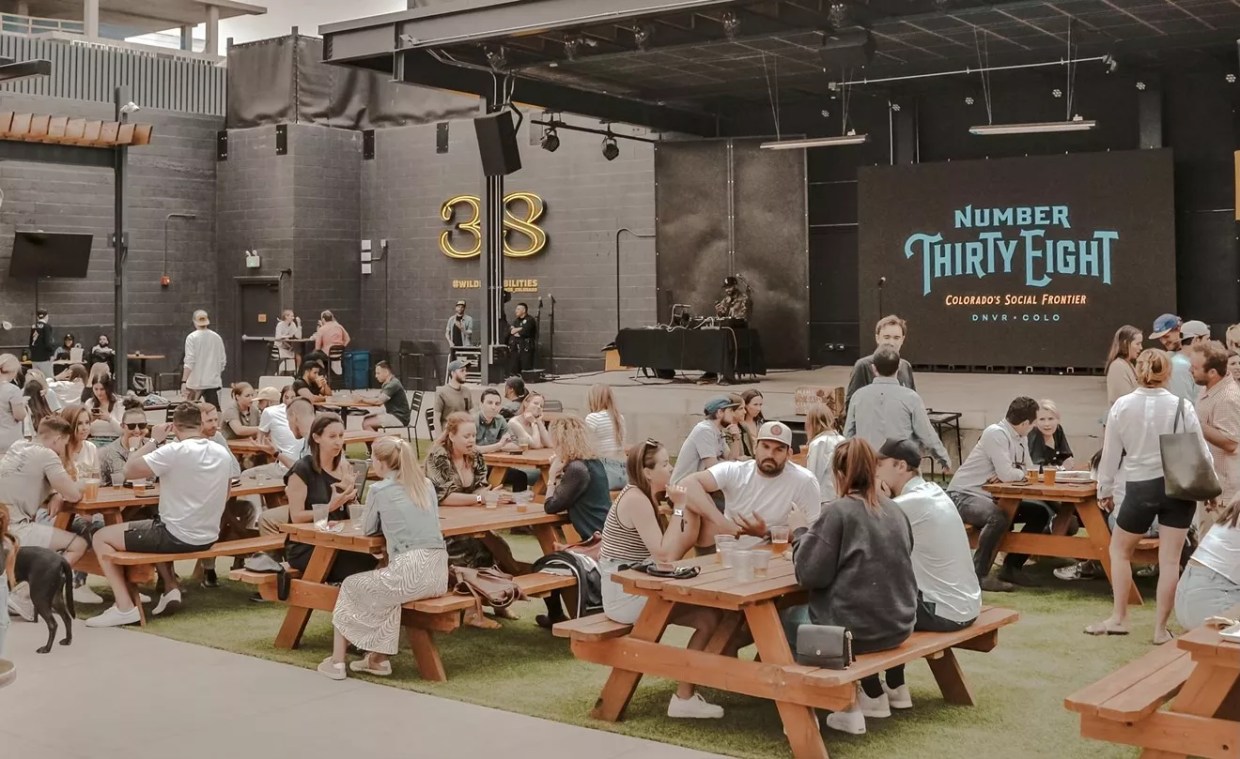 A mix of people in their mid-thirties sits at picnic tables in front of an outdoor stage displaying a projected banner with "Number Thirty Eight" in large letters.