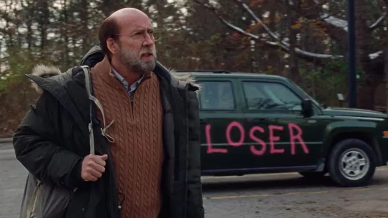man in sweater and jacket in front of a car that has "loser" written on it in graffiti