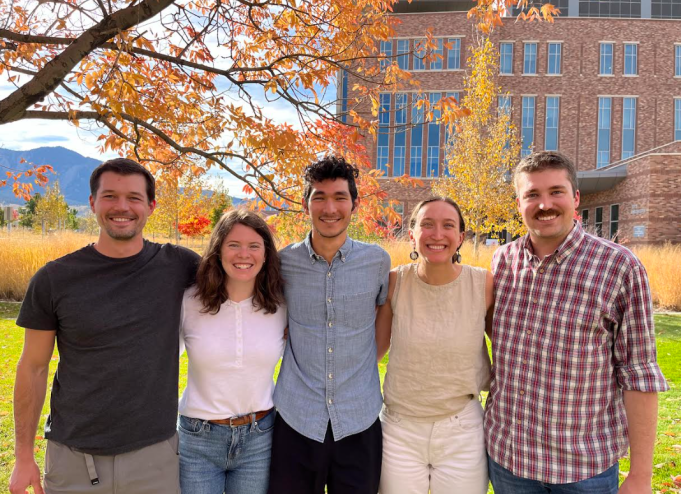 Five people smile at the camera arm in arm.