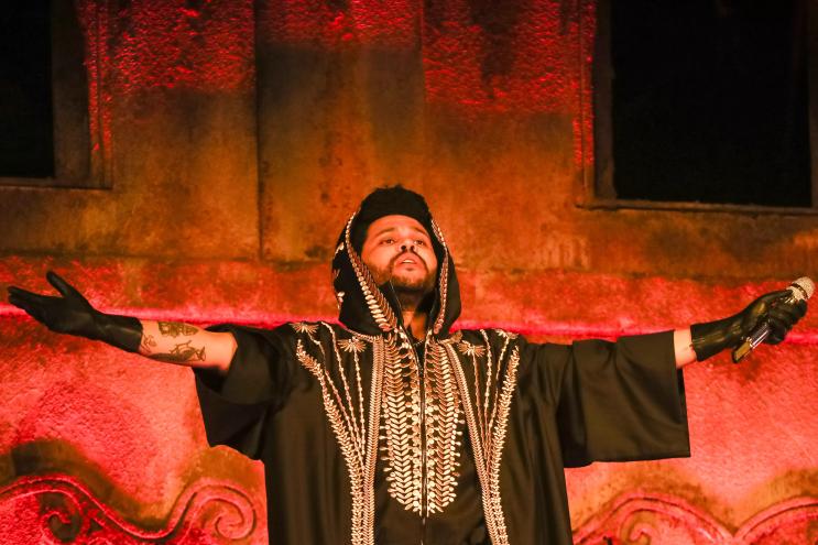 The Weeknd in front of an orange screen with his arms spread open