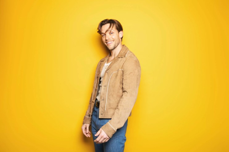Pop singer John K stands in front of a yellow background while wearing a brown jacket, blue jeans and a smile on his face.