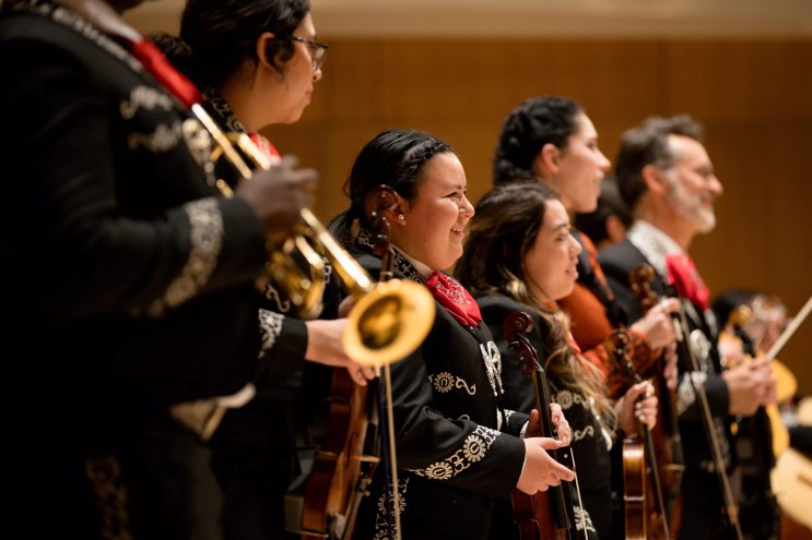 6 mariachi musicians stand on a stage while facing a crowd of supporters and fans.